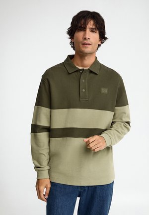 Hombre con cabello oscuro, vestido con una camisa tipo polo de manga larga a rayas verdes y beige y vaqueros azules, de pie frente a un fondo blanco.