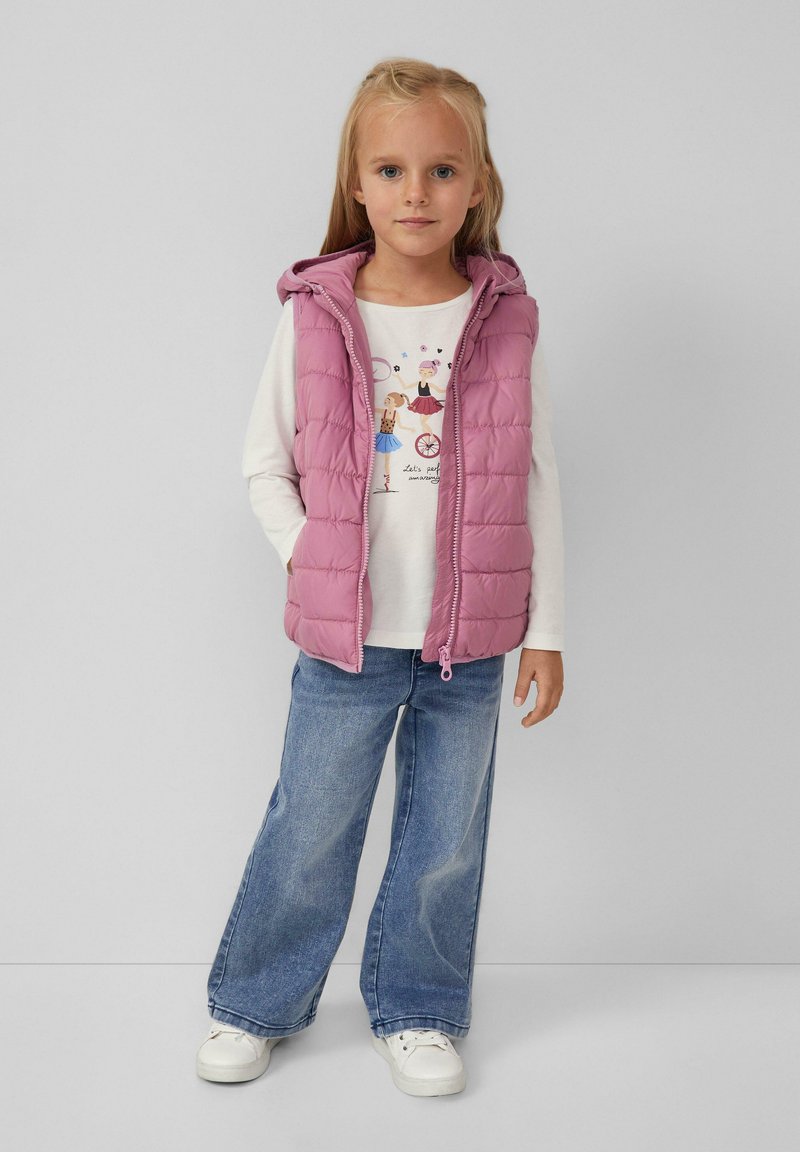 Junges Mädchen trägt eine pinke Hoodie-Weste, ein weißes Langarmshirt mit Ballerina-Druck, weite Jeans und weiße Turnschuhe vor einem grauen Hintergrund.