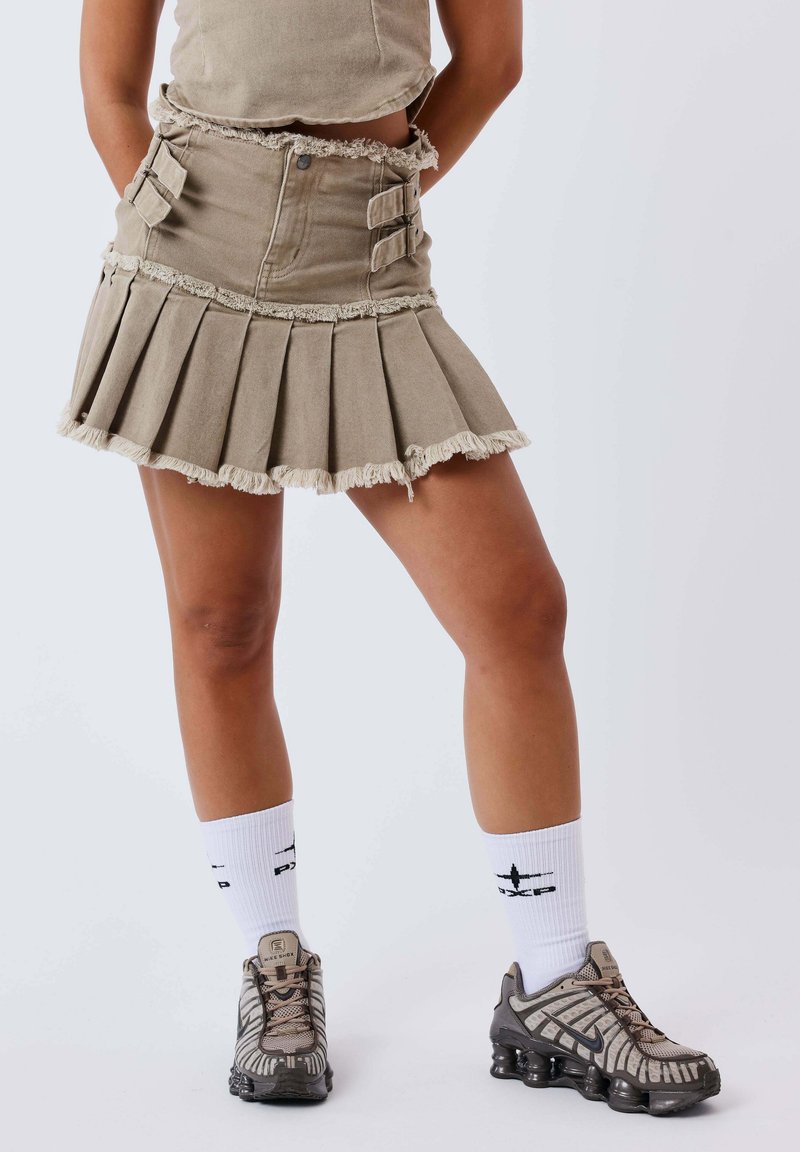 Personne portant une jupe plissée beige aux bords effilochés, des chaussettes hautes blanches et des baskets Nike grises et noires, debout devant un fond uni.