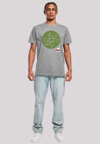 Camiseta de algodón gris con un gráfico circular verde que presenta un diseño de átomo y líneas blancas, combinada con jeans azul claros y zapatillas blancas.
