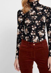 Svart blommig bodysuit med lång ärm, prydd med vita och bruna blommor, ihop med högmidjade kastanjefärgade kordbyxor med framfickor.