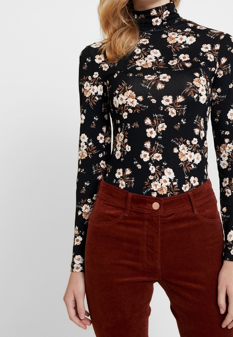 Svart blommig bodysuit med lång ärm, prydd med vita och bruna blommor, ihop med högmidjade kastanjefärgade kordbyxor med framfickor.