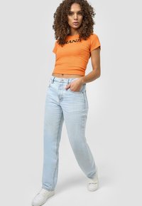 Oransje cropped t-skjorte med svart tekst, kombinert med lyseblå høytlivsjeans. Modellen har på seg hvite joggesko. Enkelt, uformelt design.