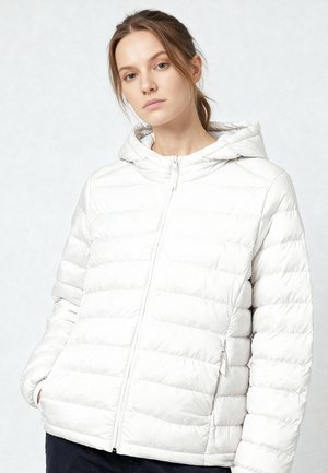 Jonge vrouw draagt een wit doorgestikt gewatteerd jack met capuchon, ritssluiting aan de voorkant en handen in de zakken, staand tegen een lichte achtergrond.