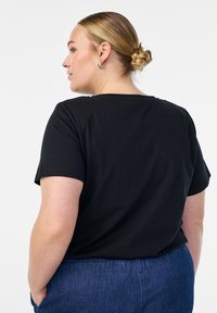 Moteris su šviesiais plaukais surištais į kuodą, dėvinti juodą trumpo rankovės marškinėlį ir mėlynus džinsus, stovi šonu su rankomis kišenėse prieš baltą foną.