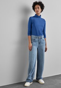 Blaues, langärmeliges Rollkragenoberteil, kombiniert mit hellblauen, weiten Jeans und weißen Sneakers. Glatte Texturen und lässige Passform.