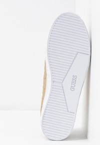Sola di sneaker bianca con scanalature testurizzate e un accento metallico dorato sul lato. Presenta il nome del marchio "GUESS" impresso in grigio.