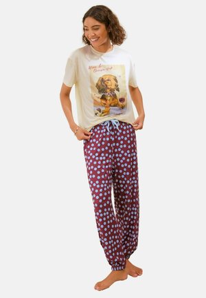 Vrouw die glimlacht, draagt een t-shirt met een teckel en tekst, gecombineerd met bordeauxrode pyjamabroek met blauwe stippen en een blauw trekkoord.