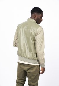 Zelená kožená bomber bunda s béžovými nylonovými rukávy, ribbovanými manžetami, texturovaným povrchem a detailem loga na zadní straně. Stylizováno s olivovými kalhotami.
