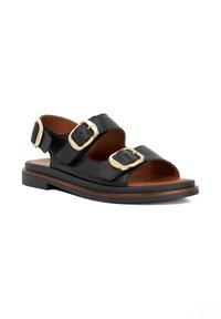 Black leather sandal featuring three adjustable straps with gold buckles, a flat brown sole, and open-toe design. Smooth texture throughout.