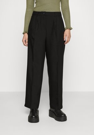 Pantalones negros de pierna ancha con pliegue frontal, cierre con botón y textura lisa, combinados con zapatos de plataforma negros y un top verde acanalado.