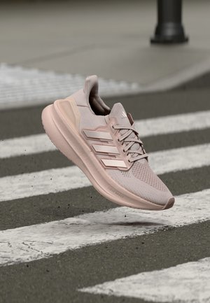 Chaussure de course Adidas rose clair avec des bandes blanches, en l'air au-dessus d'un passage piéton sur une rue en asphalte.