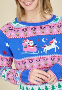 Frau, die einen bunten Weihnachtspullover trägt, auf dem der Weihnachtsmann in einem rosa Schlitten sitzt, der von einem weißen Rentier gezogen wird, umgeben von Rentieren, Sternen, Bäumen und festlichen Mustern.