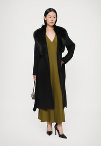 Cappotto nero con colletto in pelliccia sintetica, indossato sopra un vestito verde. La modella tiene una piccola borsa e indossa décolleté nere. Design semplice ed elegante.