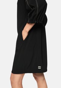 Schwarzes Kleid mit lockerem Schnitt, Puffärmeln, seitlichen Taschen und kontrastierender Naht. Mit kleinem Markenaufnäher am Saum.
