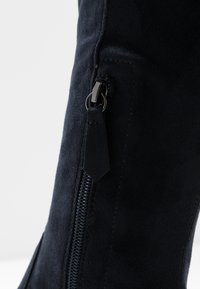 Botte noire en daim montant jusqu'au genou avec une fermeture éclair latérale ornée d'une petite étiquette en cuir. La texture est douce, et le design est élégant et ajusté.