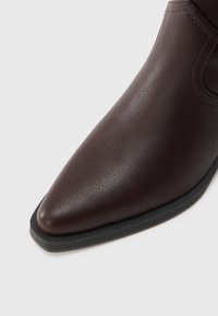 Sötétbarna bőr bokacsizma hegyes orral és texturált felülettel. Fekete gumitalppal és finom varrásdíszítéssel.