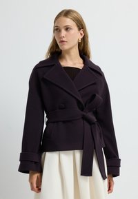 Mörk lila dubbelknäppt jacka med bred krage, bälte i midjan och stora knappar. Har strukturerade ärmar och en mjuk textur.