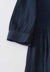 Tessuto di un blu navy, con una superficie testurizzata e un design increspato. Presenta maniche lunghe e gonfiate con una finitura a polsino e una vita arricciata.