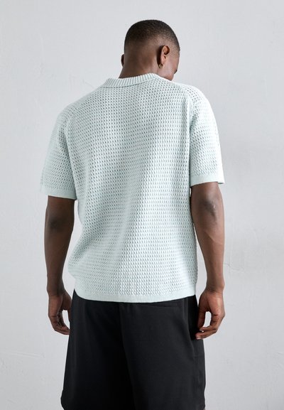 Top de punto de color azul claro, de manga corta, con un patrón texturizado y cuello acanalado, combinado con pantalones cortos negros, visto desde la parte de atrás.