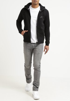 Muž kráčející v černé mikině na zip značky Nike, bílém tričku, šedých džínách a bílých teniskách na jednobarevném pozadí.