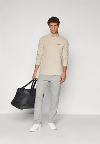 Beige sweatvest met rits en logo, grijze joggingbroek, witte sneakers en een zwarte reistas, staand tegen een neutrale achtergrond.