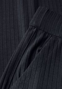 Tessuto nero a coste con una morbida texture presenta una cintura e dettagli nelle cuciture laterali. Il design include strisce verticali per un aspetto strutturato.