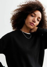 Sudadera negra de corte relajado, con textura suave y puños acanalados, combinada con un delgado collar de cadena plateada. El cabello rizado enmarcó el rostro.