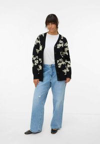 Cardigan noir avec motif floral blanc, texture côtelée, fermeture par boutons, associé à un jean bleu clair à jambes larges et des chaussures noires.