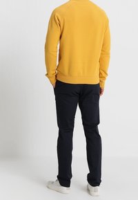 Sweat-shirt jaune avec poignets et ourlet côtelés, assorti à un pantalon bleu marine et des baskets blanches. Le tissu semble texturé, avec une coupe décontractée.