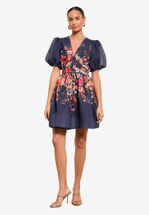 Nő sötétkék rövid ruhában, ami fodros ujjas és színes virágmintás, bézs magassarkú szandállal párosítva.