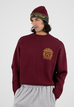 Pull bordeaux à col rond et coupe décontractée, avec un logo brodé doré. Porté avec un bonnet en tricot vert et jaune.
