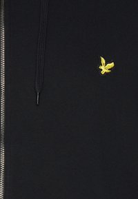 Hoodie noir à zip en mélange de coton doux. Il présente un petit logo d'oiseau jaune sur la poitrine et des cordons de serrage texturés.