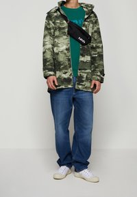 Veste imprimée camouflage en nuances de vert, portée sur une chemise turquoise, associée à un jean bleu et des baskets blanches. Sac bandoulière noir.