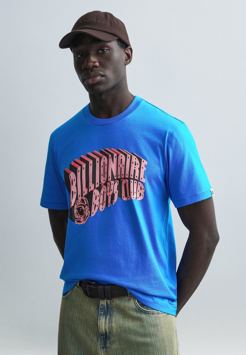 Μπλε βαμβακερό μπλουζάκι με μεγάλο ροζ κείμενο "Billionaire Boys Club". Συνδυασμένο με ξεθωριασμένο τζιν και καφέ καπέλο.