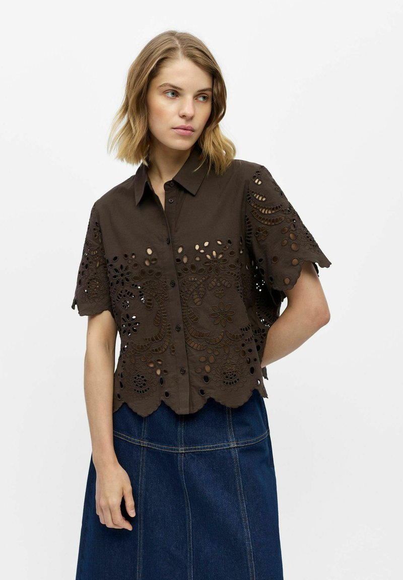 Femme portant un chemisier marron foncé à manches courtes, boutonné, avec des motifs floraux ajourés et une jupe en jean bleu foncé.