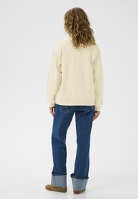 Sweat-shirt crème avec un design de fleurs brodé sur les manches, associé à un jean bleu avec des ourlets retroussés et des chaussures marron.