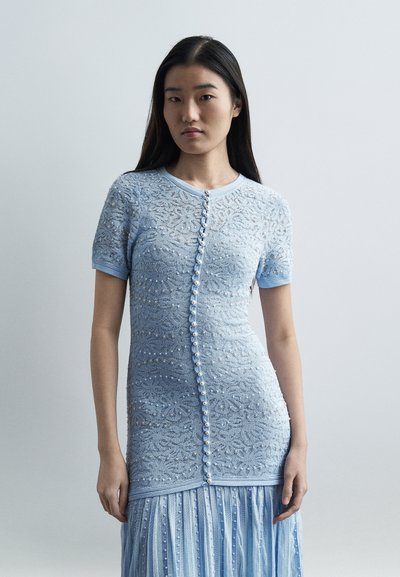 Jeune femme aux longs cheveux bruns portant une robe en dentelle bleu clair avec des boutons en perle sur le devant et une jupe plissée, sur un fond gris uni.
