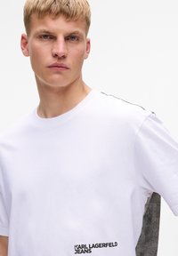 Weißes Baumwoll-T-Shirt mit schwarzem Logodruck, grauem, strukturiertem Design an der Seite und klassischem Rundhalsausschnitt. Glatte Verarbeitung.