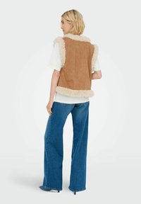 Braune Wildlederweste mit flauschigem cremefarbenem Kragen und Saum, kombiniert mit weitgeschnittenen blauen Jeans. Die Weste hat eine lockere Passform und eine weiche Textur.