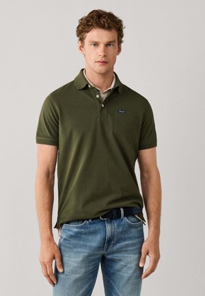 Joven con cabello rizado que lleva una camisa polo verde oliva y vaqueros azules, posando frente a un fondo claro y liso.