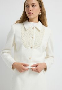 Cárdigan blanco con una tela texturizada, acentos de perlas en el pecho y cierres de botones dorados. El diseño se completa con cuello y mangas largas.