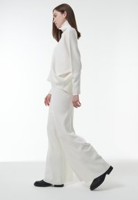 Witte coltrui en wijde broek van zachte gebreide stof. Model draagt zwarte laarzen. Gladde textuur, ontspannen silhouet, minimalistisch ontwerp.