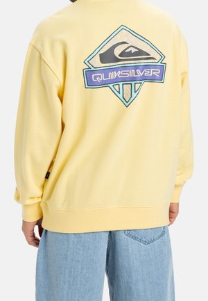 Person trägt einen gelben Pullover mit einem mehrfarbigen Quiksilver-Logo auf dem Rücken, kombiniert mit hellblauen Jeans.