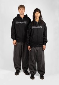Zwarte zip-up hoodies met witte grafische tekst, gecombineerd met losse donkergrijze broeken en zwarte schoenen. Glad materiaal, casual ontwerp.