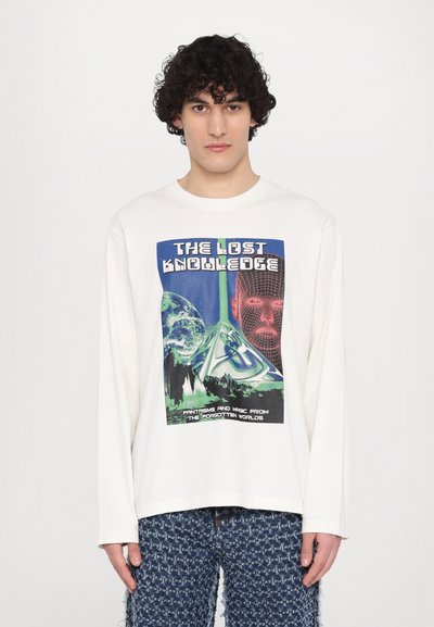 Chemise blanche à manches longues avec un imprimé graphique coloré comportant le texte "Le Savoir Perdu" et des illustrations de pyramides et d'un visage.
