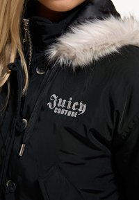 Частичен изглед на човек, носещ черно яке Juicy Couture с бяла козина по качулката и бродирано лого на гърдите.