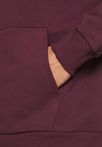 Sudadera color burdeos con un bolsillo frontal, puños y dobladillo acanalados. Textura suave con detalles de costura en contraste.