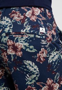 Blå blommiga byxor med ett djärvt mönster av stora rosa och gröna blommor. Inkluderar en bakficka med en etikett som detaljkerar märket.