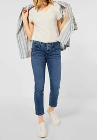 Chemise blanche à col en V texturé, jeans slim bleu, et baskets blanches. Un blazer rayé est drapé sur les épaules.